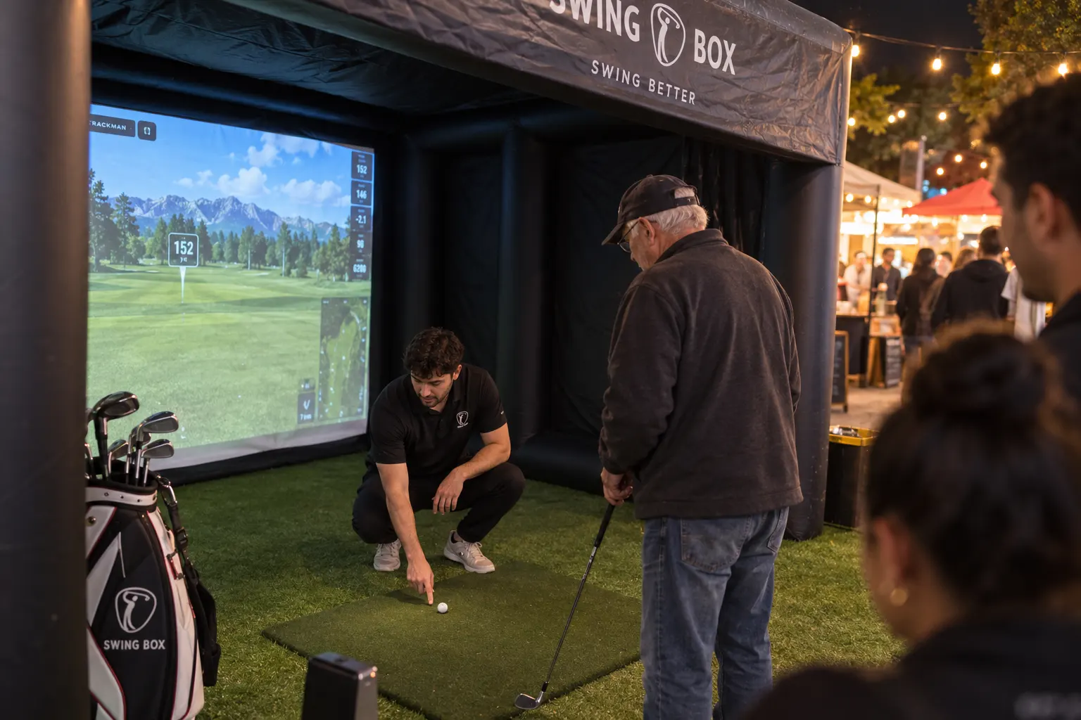 Swing Box attendant places a golf ball on the mat while a guest waits inside the black inflatable simulator at a night event
